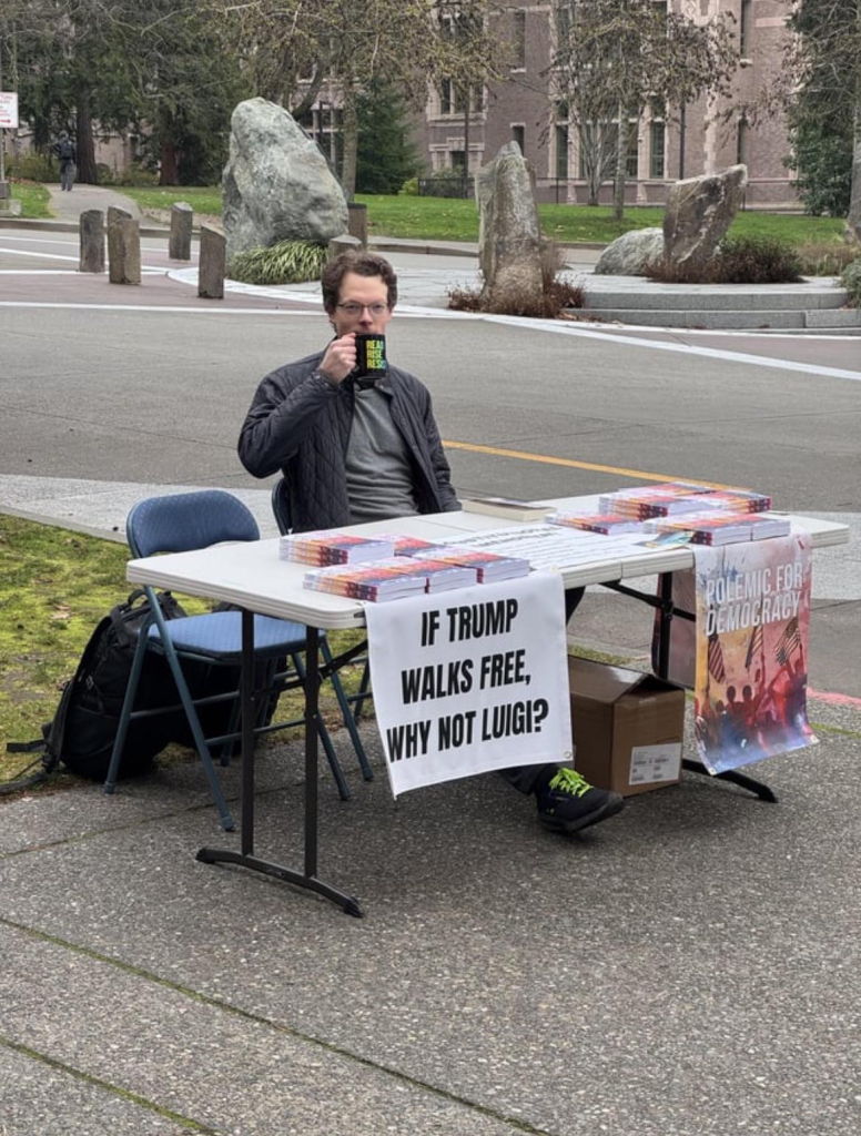 A man sipping coffee sits at a folding table outside on a sidewalk.There are various documents spread out on the table in front of him and there's a sign hanging from the table that says "If Trump walks free, why not Luigi?" in black letters on a white background.