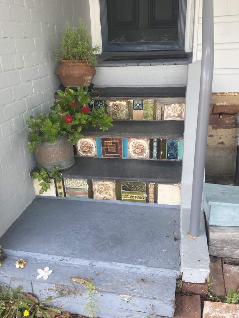 Three grey painted concrete house steps with non-matching tiles on their face. Two ceramic pots with plants rest on the left of the first and third steps. The terrace house has white painted brick and weatherboard and a dark charcoal painted door.