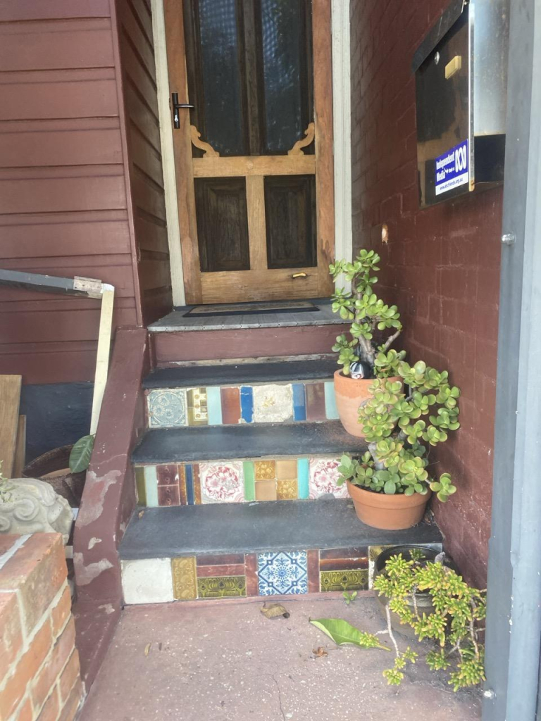 Three grey painted concrete house steps with non-matching tiles on their face. Three ceramic pots with succulent plants rest on the right of the each step. The terrace house has brown painted brick and weatherboard and a wood stained door.