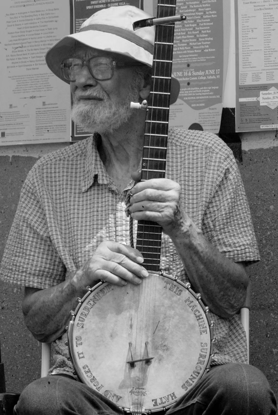 A black-and-white 2004 photograph of American folk singer-songwriter, musician and social activist Pete Seeger, an 85 year old white man with a gray beard, wearing glasses, a wide brim sun hat, and a short-sleeved shirt.

Seeger is sitting down and holding his banjo, squinting and looking into the distance. A few posters are visible on a wall behind him.

The banjo has text inscribed along its edge:

"This machine surrounds hate and forces it to surrender"