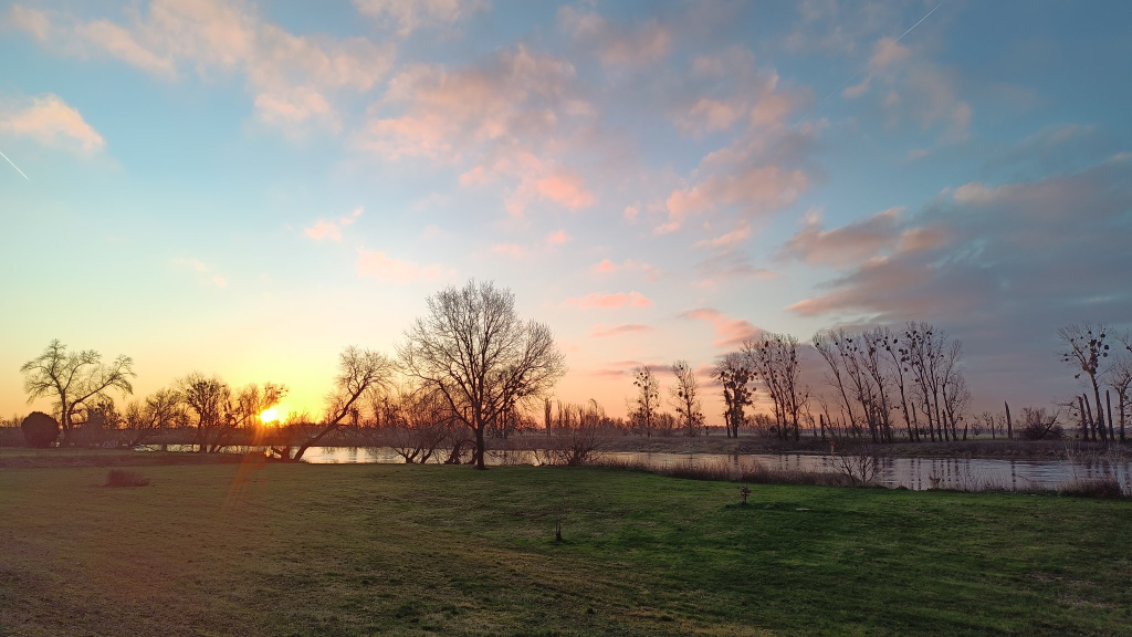 Wir schauen an der Elbe Richtung Sonnenaufgang.
Vor uns liegt eine grünlich schimmernde Wiese, dahinter folgt der Fluss Elbe, der quer durch das Bild fließt.
Am gegenüber liegenend Ufer stehen Bäume kahl am Ufer und recken ihre Äste in den Himmel
Hinter den Wiesen am gegenüber liegenden Ufer, ist die Sonne gerade aufgegangen und leuchtet rotgelb zu uns.
Am Himmel sind ein paar kleine Wolken, die von der Sonne rötlich von unten beschrieben werden