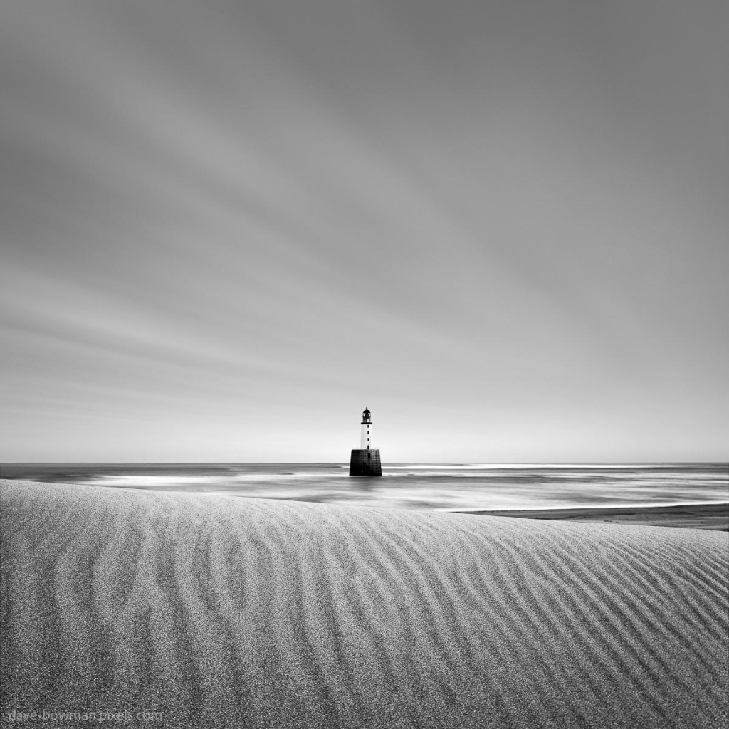 A photograph features Rattray Head Lighthouse, standing alone on the horizon in Rattray, Scotland. The long exposure creates a smooth, tranquil effect on the rippled sand in the foreground, which forms a natural path leading toward the lighthouse. Soft, streaked clouds drift across the sky, adding a sense of movement to the minimalist composition, while the interplay of light and texture brings depth to the monochrome scene.