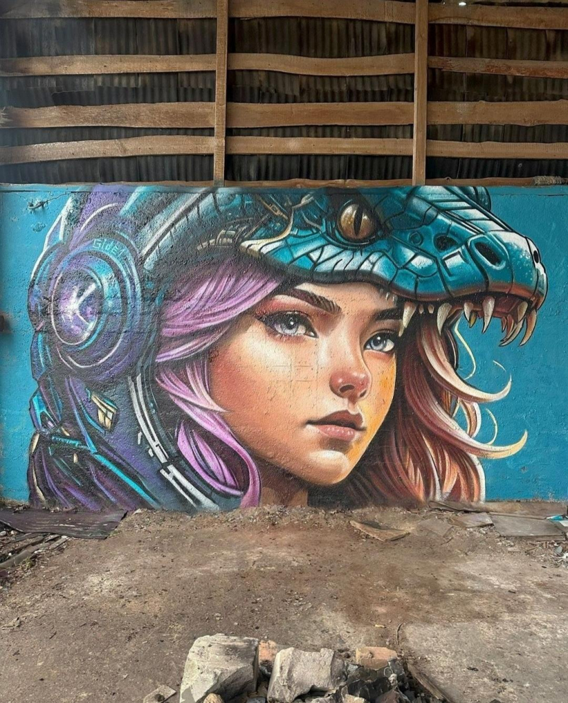 Streetartwall. A very beautiful mural of a young woman in manga style was sprayed/painted on a street wall. The woman with wavy, pink/blonde hair wears an astronaut helmet in the shape of a green snake head and looks seriously into the distance. According to the artist, the mural means that the astronaut wants to conquer new worlds and that the snake is the symbol for the year 2025. It is her talisman and is intended to help her carry out a successful expedition to unite the inhabitants of Earth with other civilizations for a common, bright future.