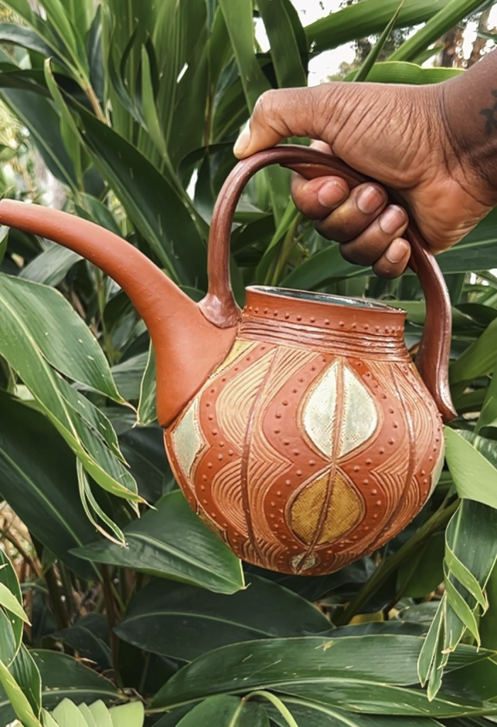 A ceramic watering can with a long spout, painted in earth tone colors with symmetrical geometric line and dot patterns, held by a brown hand in front of tropical foliage. 