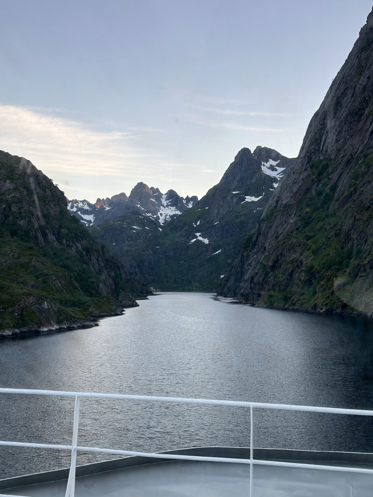 A very narrow gorge seen from the deck of a ship, with blue midnight sun sky, over steel water, and sheer rocky cliffs to port and starboard.  Snowy peaked mountains ahead.