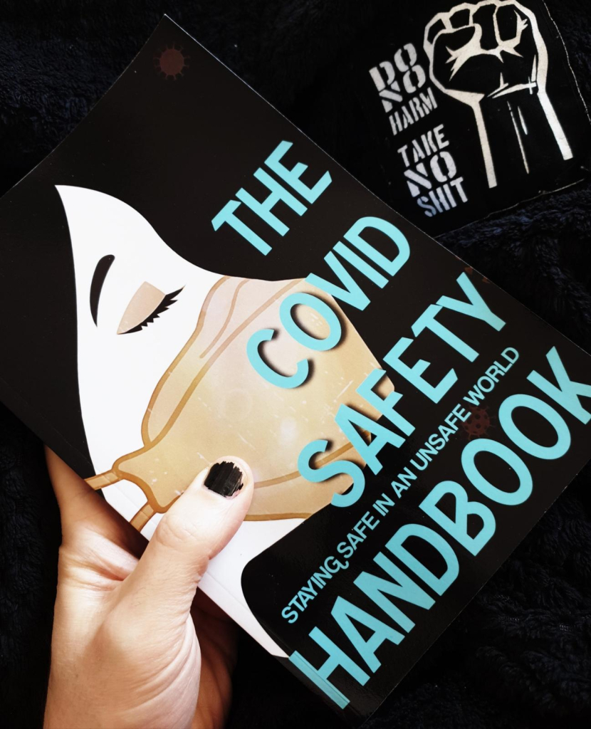 The Covid Safety Handbook: Staying Safe in an Unsafe World by Violet Blue, hardcopy format, held by a hand with chipped-painted fingernails over a blanket with a patch that reads "do no harm, take no shit."