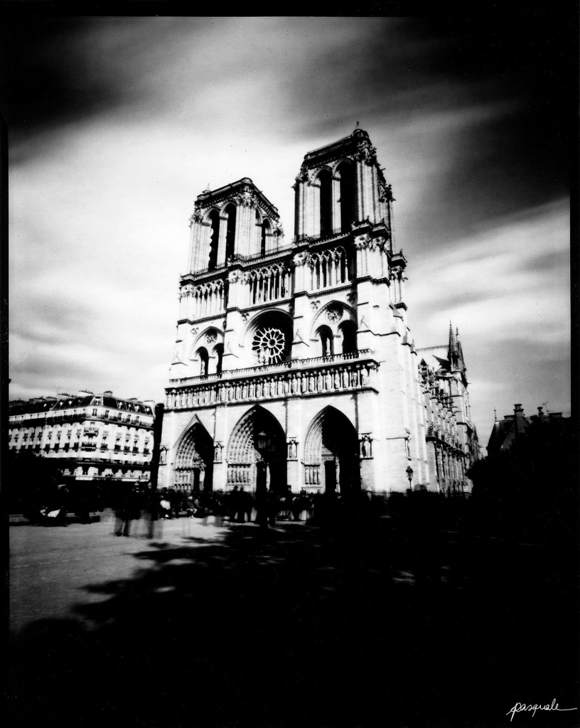 Photographie en noir et blanc représentant la façade de Notre-Dame-de-Paris prise de côté. Le temps de pause étant assez long, les nuages et la foule forme un filet sombre.