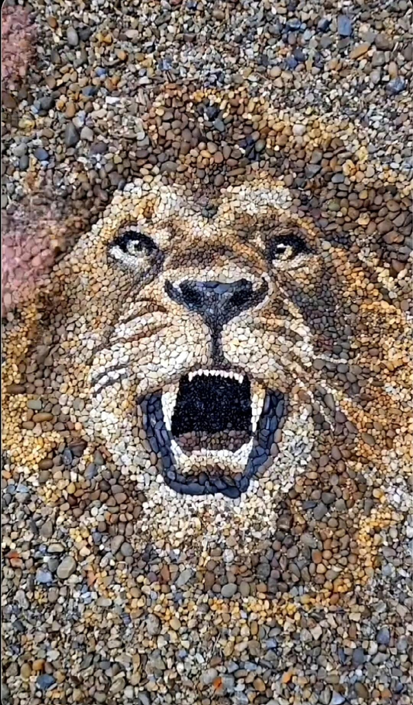 Urbanart. An impressive mosaic of small gray, brown, black and yellow stones. It is the head of a lion with its mouth open. It has been placed as a mosaic image between stones on the beach and looks a little menacing.