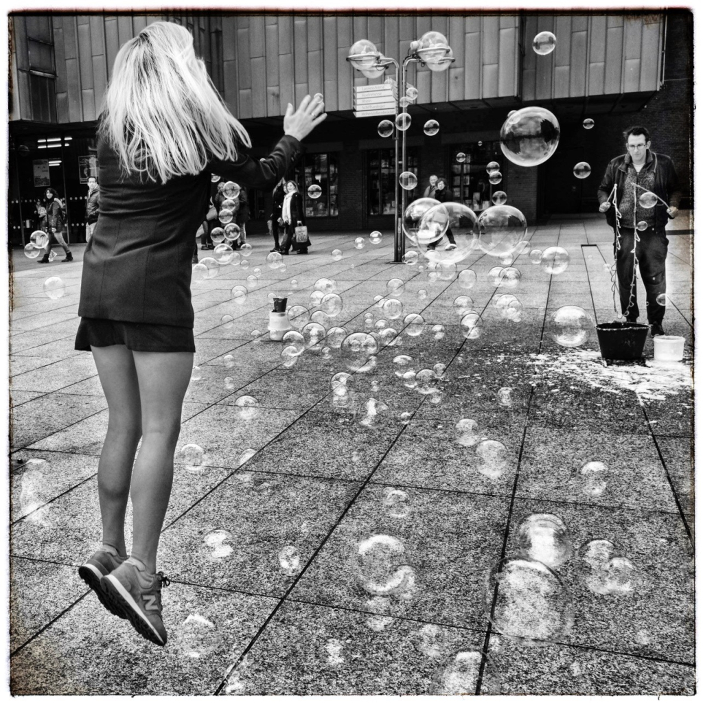 On the paved forecourt of a city museum a blonde woman in sneakers and shorts jumps in the air among a cloud of large soap bubbles. The bubble artist stands on the right in the background. 