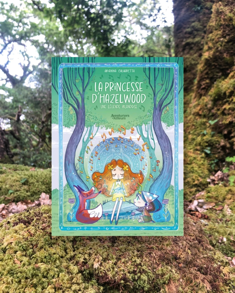 Livre posé sur de la mousse en forêt. La couverture de La princesse d’Hazelwood montre une jeune fille assise entre deux arbres au côtés d’un autres jeune fille et d’un renard, tenant un artéfact produisant de la musique.