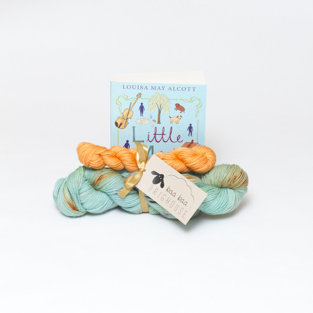 Picture shows a sock set of yarn consisting of a main skein which is aqua with orange, purple and green speckles and a mini skein which is orange. They are tied together with gold ribbon. Behind the yarn stood on its end is a copy of Little Men by Louisa May Alcott. The book is aqua with purple and green drawn images on it.