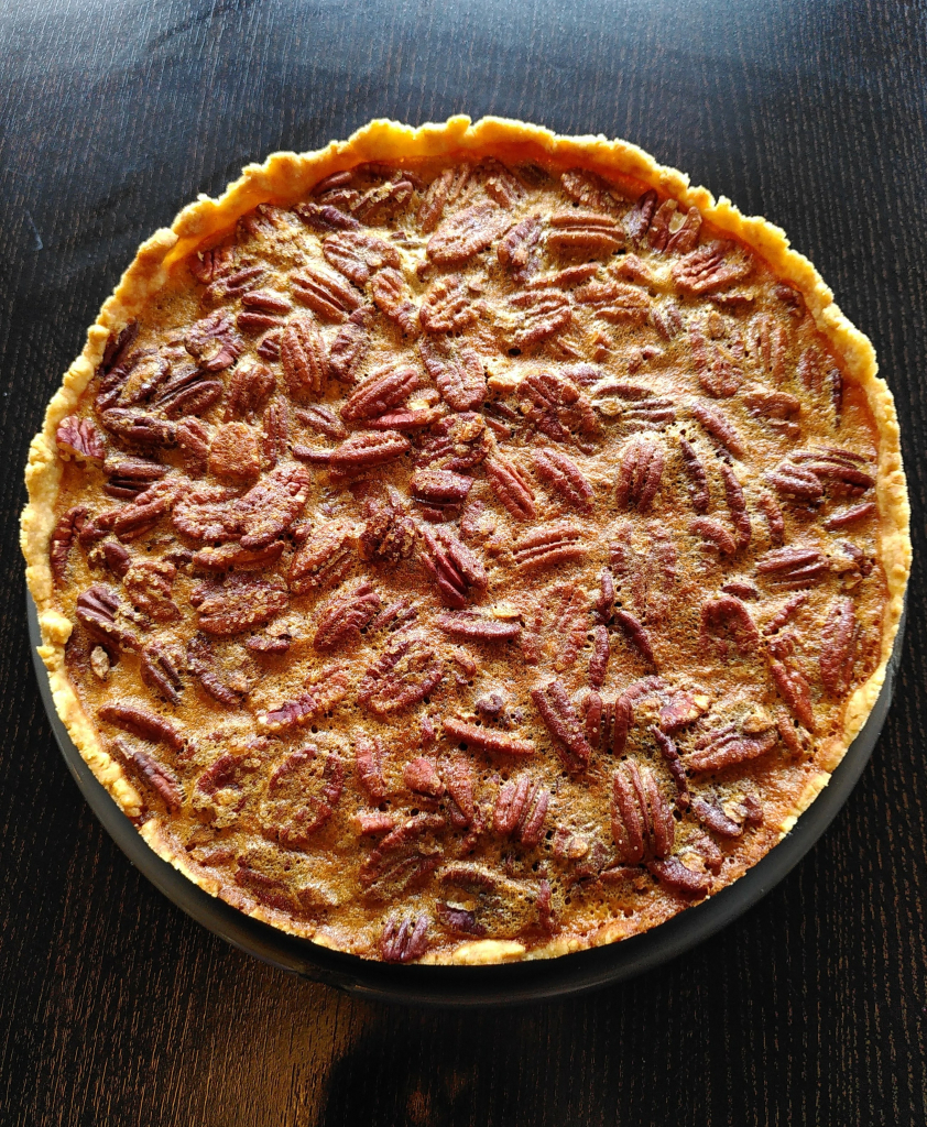 Ein runder Maple Pecan Pie. Dabei handelt es sich um einen buttrigen Mürbeteigboden, der eine Füllung aus vielen Pekannüssen und eine Crème mit Ahornsirup hat. Viele der braunen Pekannüsse sind an der Oberfläche zu sehen, die gebackene Füllung hält die Nüsse an Ort und Stelle. Sie glänzt goldgelb.