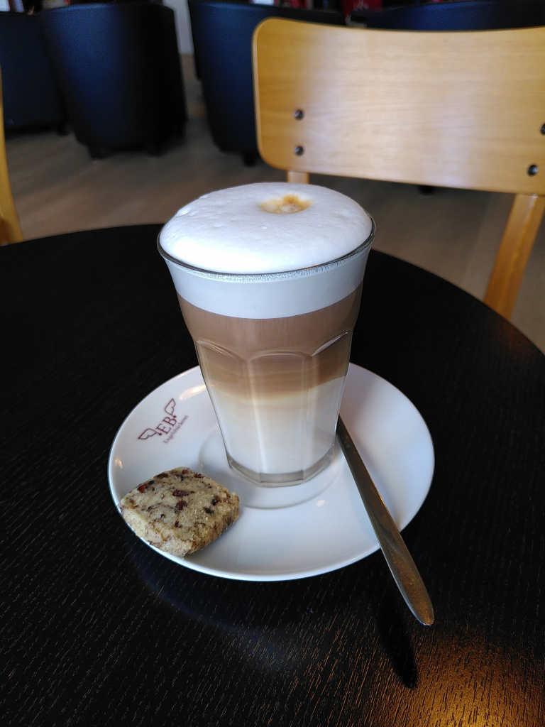 Auf einem dunkelbraunen Holztisch steht eine weiße Untertasse mit einem Glas Latte Macchiato. Links neben dem Glas liegt ein selbstgebackenes, viereckiges Plätzchen, rechts ein langstieliger Löffel zum umrühren. 
Ein Latte Macchiato bedeutet über setzt *gefleckte Milch* und  besteht aus viel heißer, geschäumter Milch (selbstverständlich geht auch pflanzliche Milch) und einem Espresso. Die Milch wird zuerst ins Glas gegeben. Der entstandene Schaum bleibt an der Oberfläche, die heiße Milch sinkt nach unten. Nun wird der frisch zubereitete Espresso in einem dünnen Strahl in die Mitte gegossen. Durch den Temperaturunterschied zwischen Schaum und Flüssigkeit setzt sich der Espresso zwischen den Schaum und die Milch. Dadurch entstehen drei Schichten: weiß, braun, weiß = Schaum, Espresso, heiße Milch.