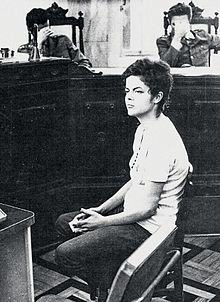 Black and white photo of Dilma Rousseff at her military court hearing. The judges in the background are hiding their faces with their hands. 