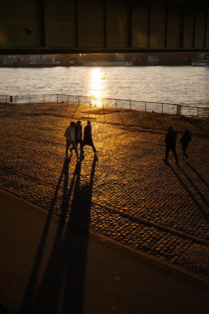 Spaziergänger gehen über ein Kopfsteinpflaster am Rheinufer in Köln, durch die tiefstehende Abendsonne werfen sie sehr lange Schatten auf den Boden. Alles wird in goldenes Licht getaucht, über den Personen ist ein Stahlträger einer Brücke zu erkennen.
Im Hintergrund der Rhein sowie ein Geländer am Ufer. 