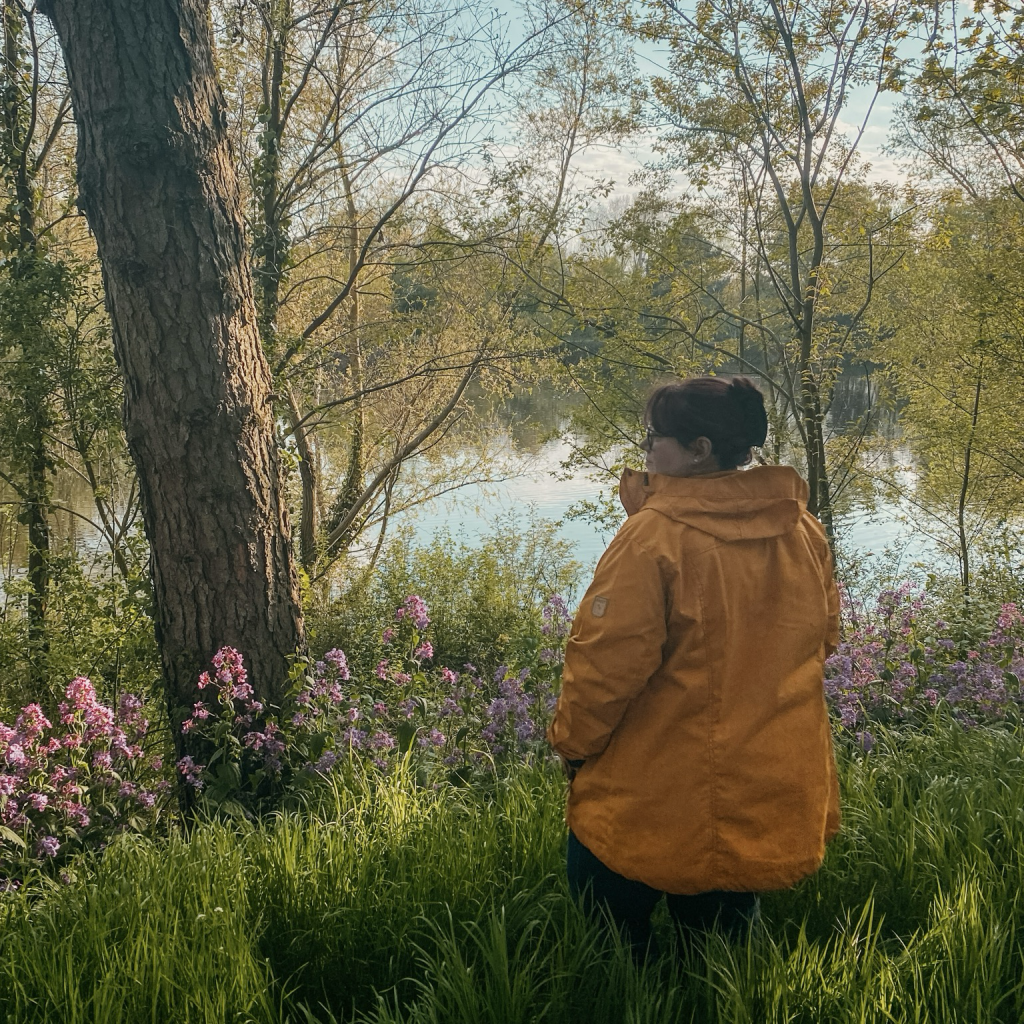 Die Post-Erstellerin steht in einer gelben Jacke auf einer Wiese am See. 