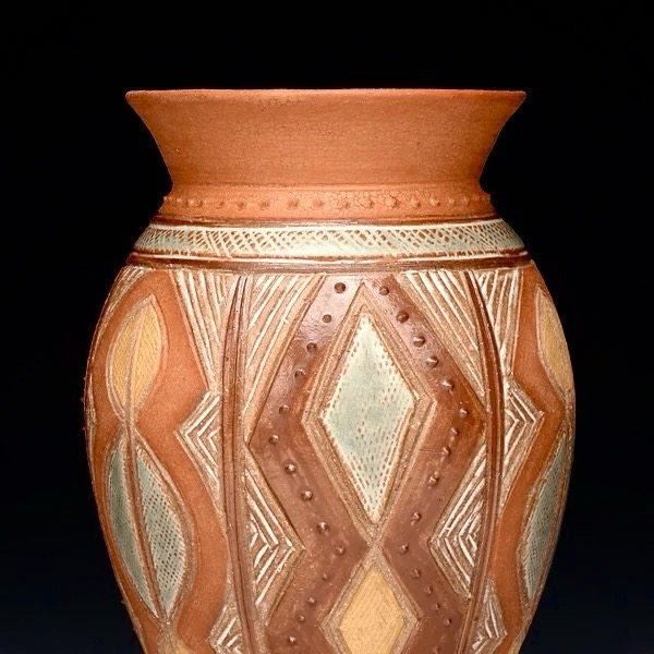 A closeup of the upper half of a vase I made. The neck flares out at an angle and is painted with the brownish-red crackle clay. The vase is decorated with geometric patterns, diamonds, lines and dots. The other colors are shiny brown, light yellow and light blue-green.  The photo is a studio lit with a plain, black background. 