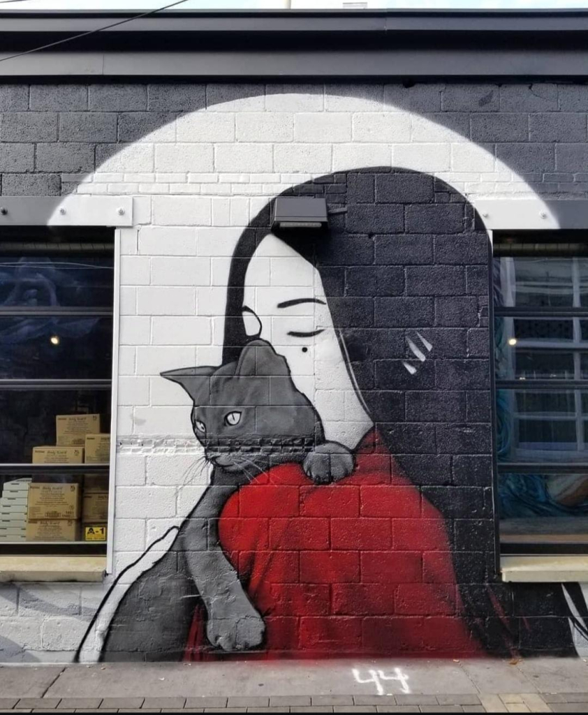 Streetartwall. A beautiful mural of a young Asian woman and a black cat has been painted on a white brick wall at the back of a small brewery. The mural is located between two windows. The black-haired woman with the pale face in a red kimono can be seen from behind. A black cat is sitting on her shoulder and she is nestles her face against the animal. Beautifully simple, but skillfully painted and designed only in black, white and red.