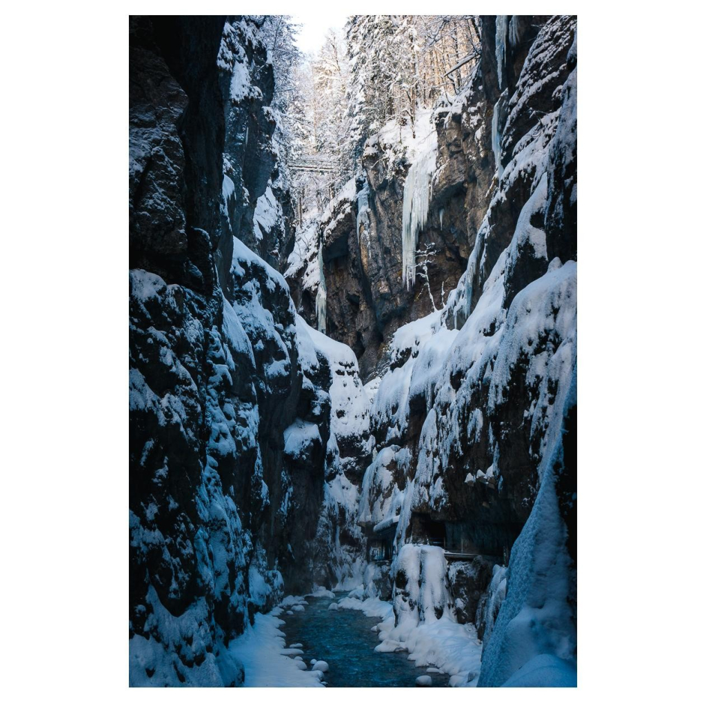 Eine tief in den Stein gegrabenes Flussbett. Auf den senkrecht nach oben ragenden Felswänden liegt Schnee. In der Ferne überspannt eine kleine Brücke die Klamm.