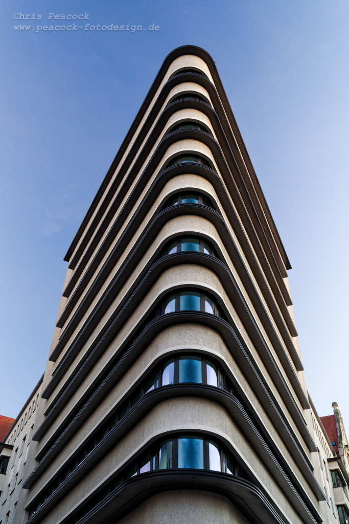 EN: The photo shows the triangular building viewed from the very narrow front side against a blue sky. With a little imagination, the arched windows on each floor look like eyes because of the round corner. The façade is light in colour and offers a nice contrast with dark edges running around each floor.

DE: Das Foto zeigt das dreieckförmige Gebäude von der sehr schmalen Stirnseite aus betrachtet vor blauen Himmel. Die wegen der runden Ecke gewölbten Fenster in jeder Etage wirken mit etwas Fantasie wie Augen. Die Fassade ist hell gefärbt und bietet mit dunklen umlaufen Kanten auf jeder Etage einen schönen Kontrast.