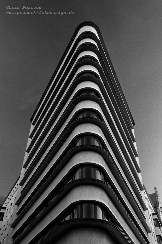 EN: The black and white edited version of the photo shows the triangular building viewed from the very narrow front side against a blue sky. With a little imagination, the arched windows on each floor look like eyes because of the round corner. The façade is light in colour and offers a nice contrast with dark edges running around each floor.

DE: Die schwarz-weiß bearbeitete Version des Foto zeigt das dreieckförmige Gebäude von der sehr schmalen Stirnseite aus betrachtet vor blauen Himmel. Die wegen der runden Ecke gewölbten Fenster in jeder Etage wirken mit etwas Fantasie wie Augen. Die Fassade ist hell gefärbt und bietet mit dunklen umlaufen Kanten auf jeder Etage einen schönen Kontrast.