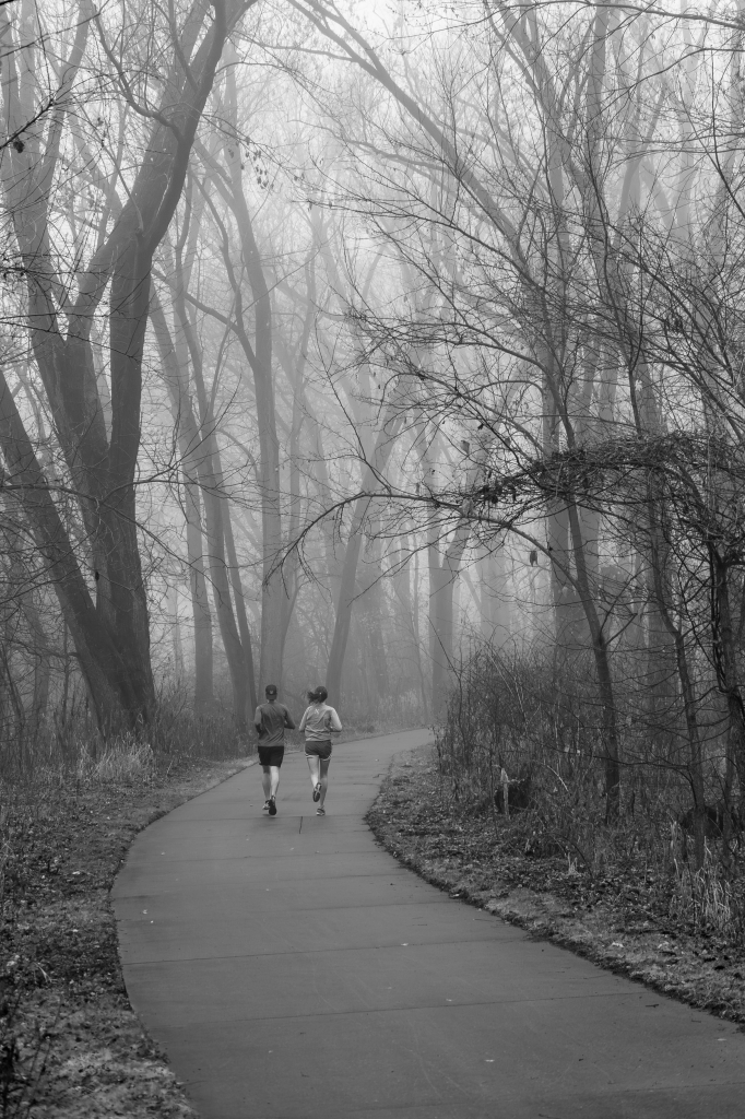 Black and white photograph of two people jogging down a paved trail winding its way through fog-shrouded, leafless trees. The trees are blackened by the moisture while still being obscured by the fog. The paved trail begins in the lower center-right frame, curves to the left, and then curves back to the right where it disappears around a corner. The joggers are dressed in shorts, long-sleeve shirts, and baseball caps.