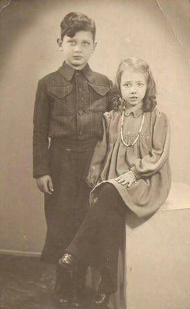 Two children posing for a vintage portrait. One child is standing, wearing a buttoned jacket and trousers, and the other child is seated, wearing a dress and necklace.