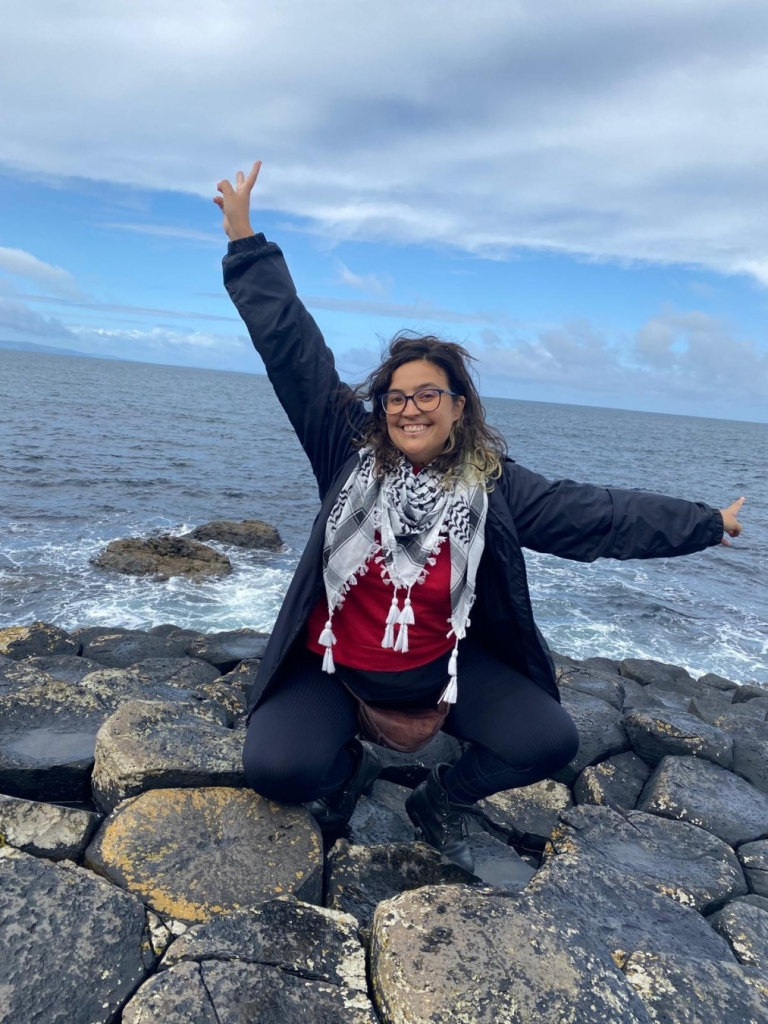 Yo en la Calzada de los Gigantes, en Irlanda del Norte, con el agua del mar de fondo. Estoy de cuclillas, sonriendo, con los brazos en forma de L.