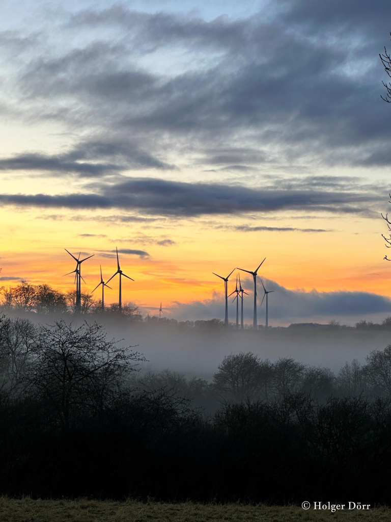 A misty landscape at sunset featuring several wind turbines silhouetted against a colorful sky with clouds.