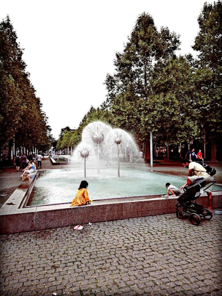 Einkaufsmeile Prager Straße in #Dresden #Pusteblumenbrunnen 

Im Vordergrund der an dieser Stelle der Einkaufsmeile gepflasterte Boden. Ein Kinderwagen steht vor der Einfassung des Brunnenbeckens. Ein kleines Kind hockt auf der Einfassung, scheint mit einer Hand nach vorn ins Wasser zu greifen, mit der anderen stützt es sich ab. Ein erwachsener Mensch - ein Bein auf dem Pflaster, eins auf der Einfassung - hält seine Hand so am Rücken des Kindes, dass er jeden Moment zugreifen könnte.

Ein paar Meter weiter sitzt ein Kind in einem gelben Kleid auf der Einfassung und schaut auf's Wasser. Die Badelatschen stehen auf dem Pflaster.

Im Hintergrund stehen Bäume, der Himmel wirkt milchig trüb.