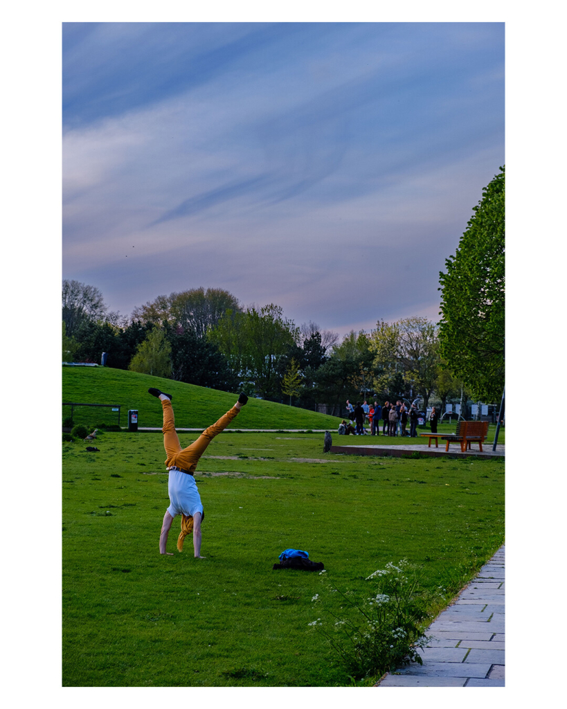 Ein grüner Park im Frühling, eine Person mit orange-brauner Hose, passendem Schal und weißem Short macht einen Handstand auf der Wiese. Die Beine sind dabei leicht gespreizt und der Schal verdeckt das Gesicht. Der Himmel ist voll mit Streifenwolken. 