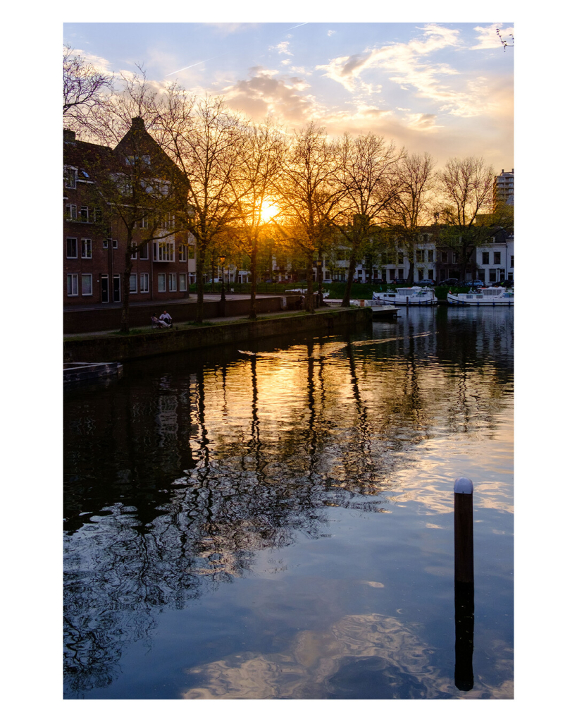 Hochkantes Foto. Ein Kanal in Utrecht bei Sonnenuntergang. Auf der gegenüberliegenden Seite des Kanals sind Hauser und Bäume ohne Blätter, durch die die untergehende Sonne scheint. Dies alles spiegelt sich im Wasser. Am Ufer sitzen zwei Personen auf Liegestühlen direkt am Wasser. 