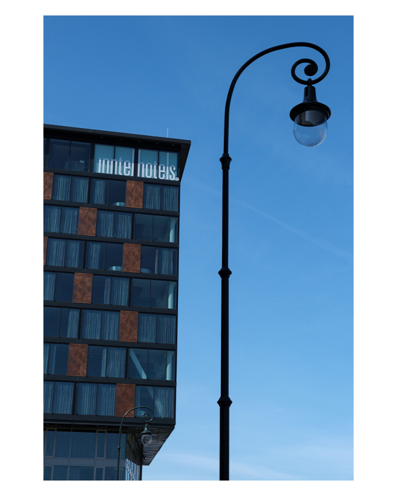 Hochkantes Bild. Eine Straßenlaterne die oben zu einem Schnörkel zusammenläuft, an diesem hängt die Lampe. Parallel zum Laternenpfahl ist ein modernes Hotel, mit vielen rechteckigen Fenstern, teils sind braune Rechtecke dazwischen. Alles ist vor strahlend blauem Himmel.