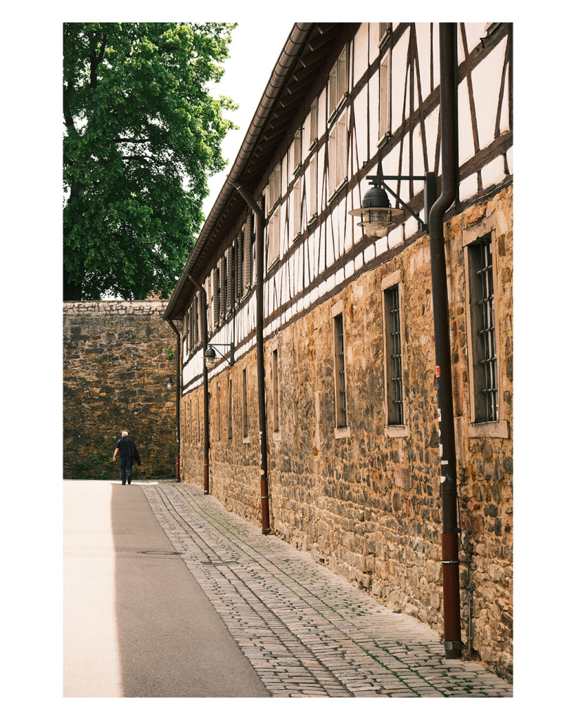 Hochlandes Bild, eine Gasse, rechts ist ein altes langes Fachwerkhaus, die untere Hälfte ist aus Stein, die obere klassisches Fachwerk, weißer Stein mit braunen Holzbalken. Am Fachwerkhaus sind Straßenlaternen angebracht. Das Gebäude wirft Schatten auf die Gasse, nur am linken Bildrand ist ein Streifen Sonne. Am Ende der Gasse ist eine hohe Mauer, eine Person läuft darauf zu. 