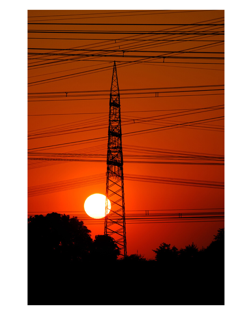 Hochkantes Foto. Leuchtend Gold-orange-roter Himmel, die Sonne ist am untergehen und steht tief am Horizont. Davor die Silhouette eines Hochspannungsmastens und Unmengen von Stromleitungen. Der untere Teil des Bildes ist die Silhouette von Bäumen. 