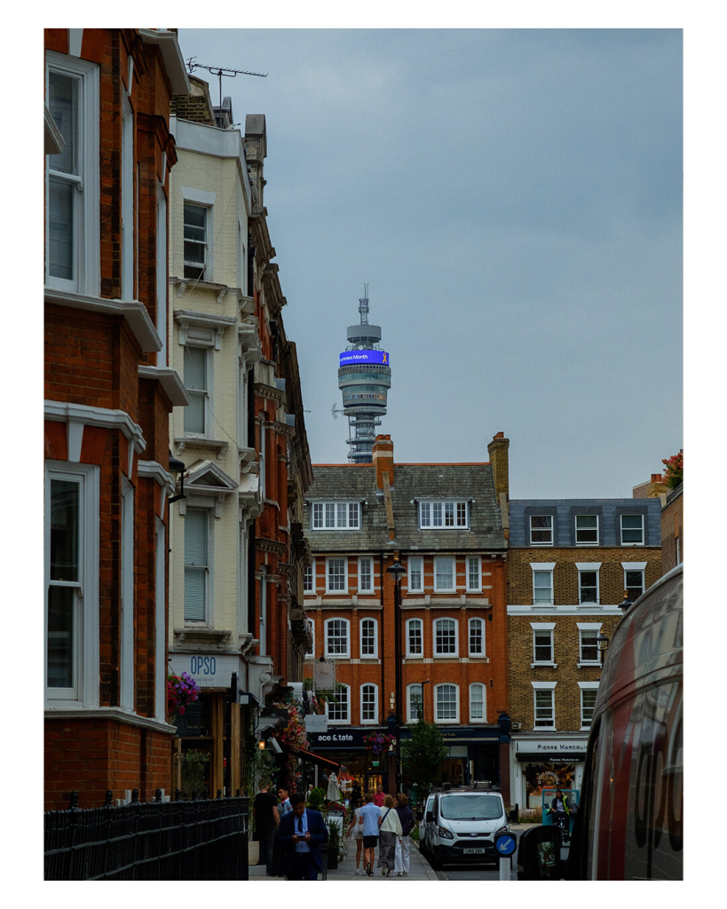 Hochkantiges Bild, eine Straße in London mit alten, gut erhaltenen Gebäuden. Auf dem Gehweg laufen Menschen. Am Horizont hinter den Gebäuden ist die Spitze des BT Towers, dem Londoner Fernsehturm. 