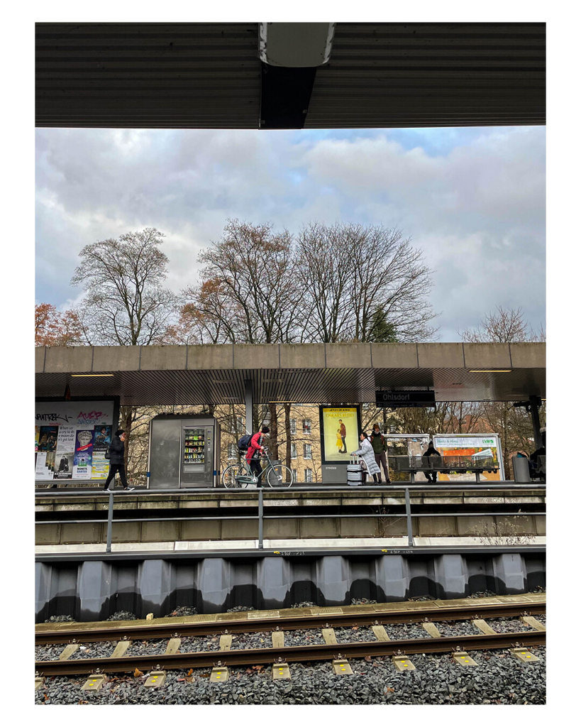 Hochkantes Bild, Blick aus einer U-Bahn auf die Haltestelle. Bahnsteig, Geländer, Dach über dem Bahnsteig, Himmel, Dach, welches oben ins Bild ragt sind alle parallel und gerade im Bild. Dadurch ist das Bild in verschiedene Teile aufgeteilt. Nur unten im Bild sind Gleise, die von rechts nach links leicht bergab gehen. 