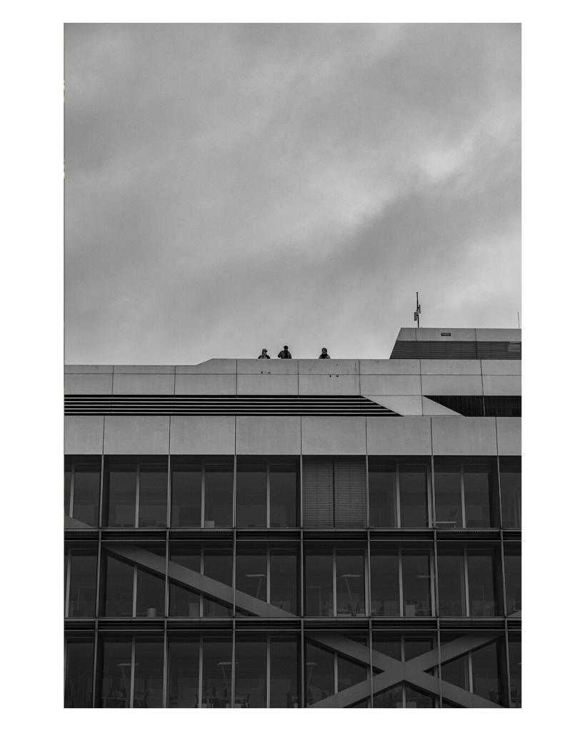 Hochkantes schwarzweiß Bild. Blick von unten Richtung Dach eines Hochhauses. Die untere Hälfte des Bildes ist das Haus, verglaste Wand, innen sind Büros zu erkennen. Auf dem Dach stehen hinter einer Mauer drei Personen, die nach unten schauen. 