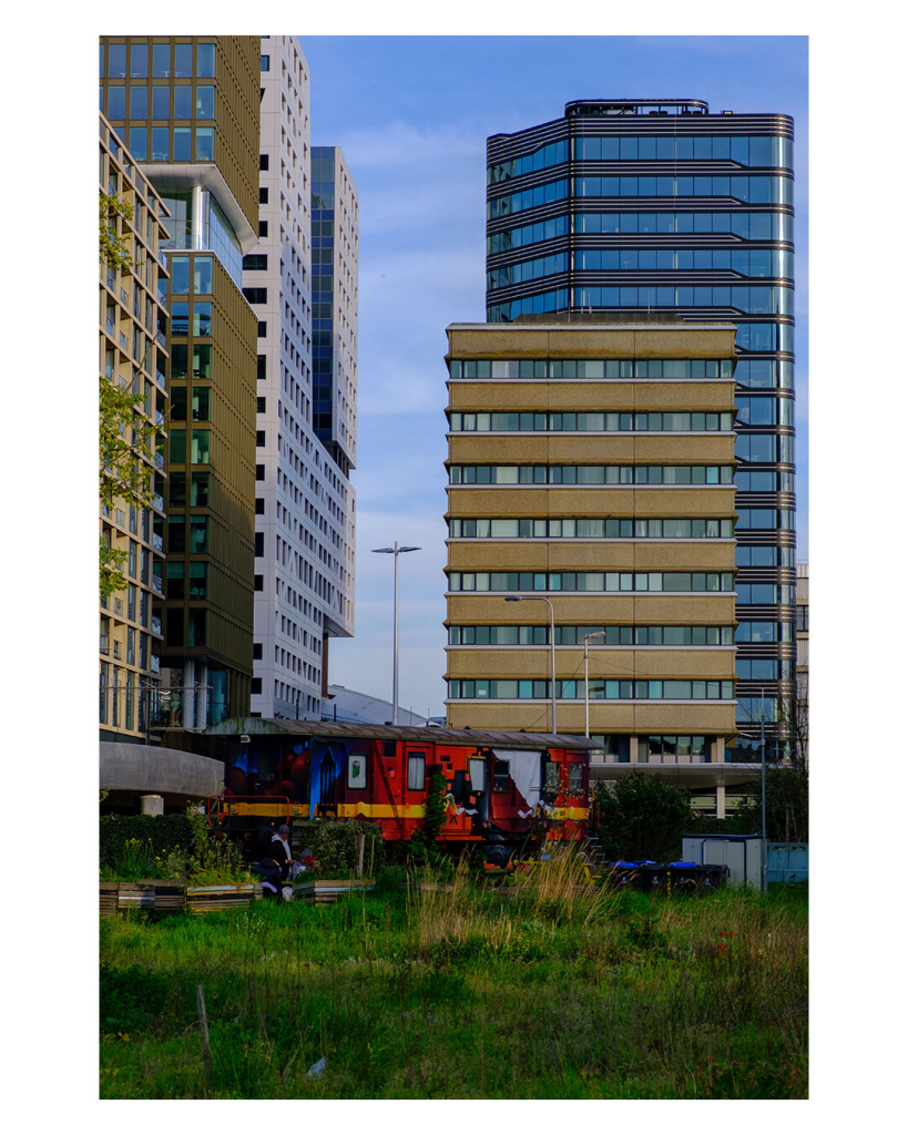 Hochkantes Foto. Im Hintergrund sind mehrere Hochhäuser des Finanzviertels in Utrecht. Davor, auf einem Stück Wiese steht ein bunt bemalter Bauwagen, vor dem ein paar Menschen sitzen, die vermutlich dort leben. 