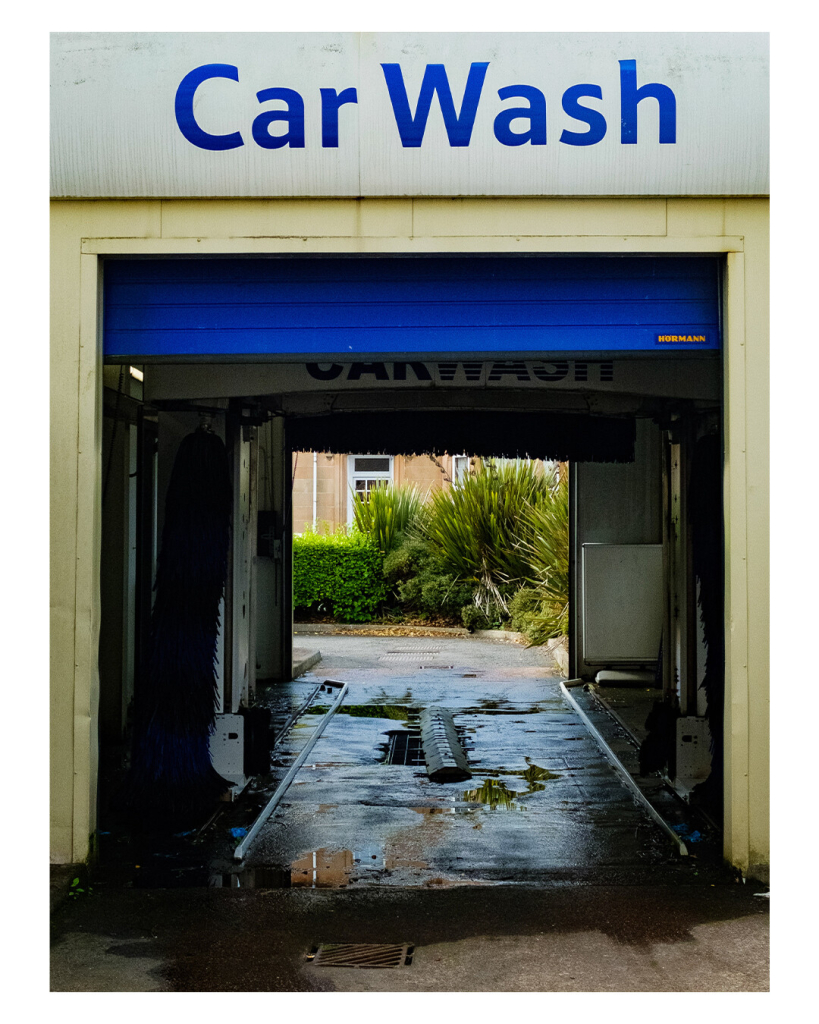 Hochkantes Foto. Blick durch eine gelblich-weiße  Autowaschanlage. Auf einem weißen Schild steht in blauen Buchstaben Car Wash, das Schild hängt über der Einfahrt. Im der Einfahrt ist oben Teil eines blauen Tors. Der Boden der Waschanlage ist voller Pfützen. Auf der anderen Seite sind Wohnhäuser und Palmenpflanzen. 