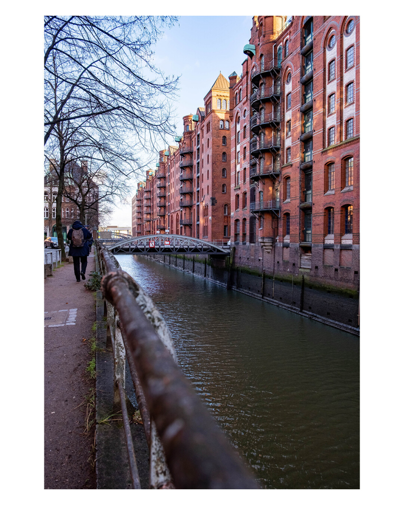 Hochkantes Bild. Rechts die Backsteingebäude der Hamburger Speicherstadt. Links davor ein Kanal über den weiter hinten auch eine Brücke verläuft. Links ist der Gehweg, der vom Kanal durch ein Geländer abgetrennt ist. Dieses geht von unten aus dem Bild heraus. 