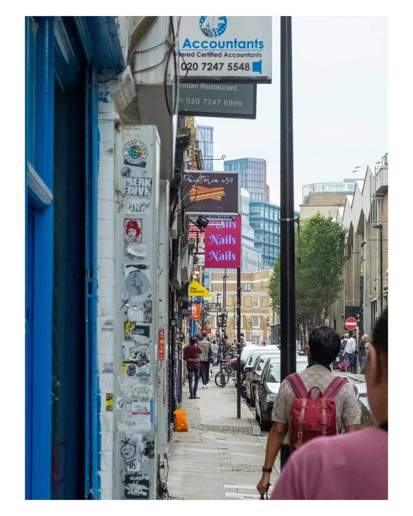 Hochkantes Foto, eine Straße in London. Links sind viele kleine Läden, viele haben ein Schild zu Straße raus hängen. Vorne ist direkt eine Wand, auf der viele Aufkleber sind. Auf dem Gehweg laufen Menschen. Die Gegend sieht eher heruntergekommen aus. Am Horizont im Hintergrund sind die Wolkenkratzer des Finanzviertels. 