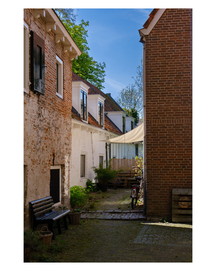 Foto im Hochformat. Blick in einen Hinterhof. Zwischen mehreren Backsteinhäusern steht eine Bank, ein Sonnenschirm und ein paar Pflanzen. An einer Wand lehnt ein Fahrrad. 