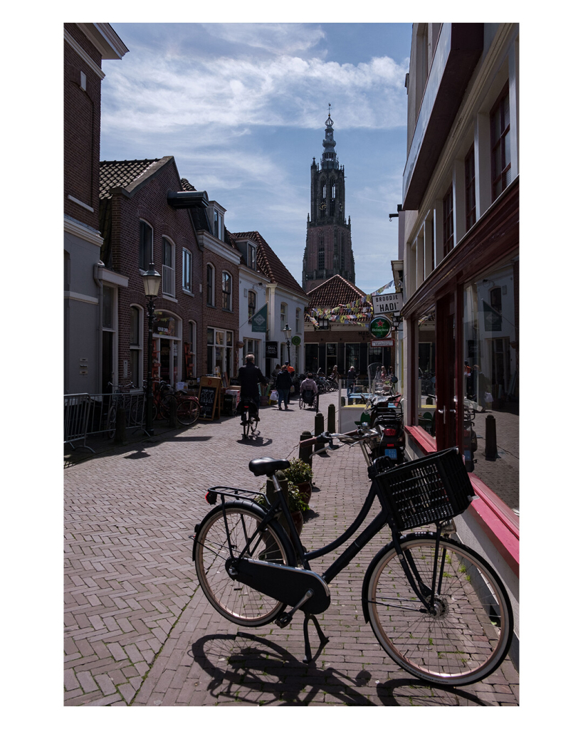 Foto im Hochformat. Eine Fußgängerzone in Amersfoort. Am Ende steht ein hoher Kirchturm. In der Fußgängerzone laufen ein paar Menschen, ganz vorne im Bild, direkt vor der Kamera ist ein abgestelltes Hollandrad. 