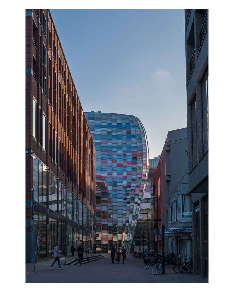 Foto im Hochformat. Eine Fußgängerzone in Utrecht. Links ist eine hohes rotes Backsteingebäude mit hohen rechteckigen Fenstern. Am Ende der Straße ist ein futuristisches rundes Gebäude, verkleidet mit vielen unterschiedlich großen rechteckigen Platten in unterschiedlichen Farben. Überwiegend türkis, hellblau und rosa. In der Fußgängerzone sind ein paar Menschen unterwegs und rechts stehen viele Fahrräder. 