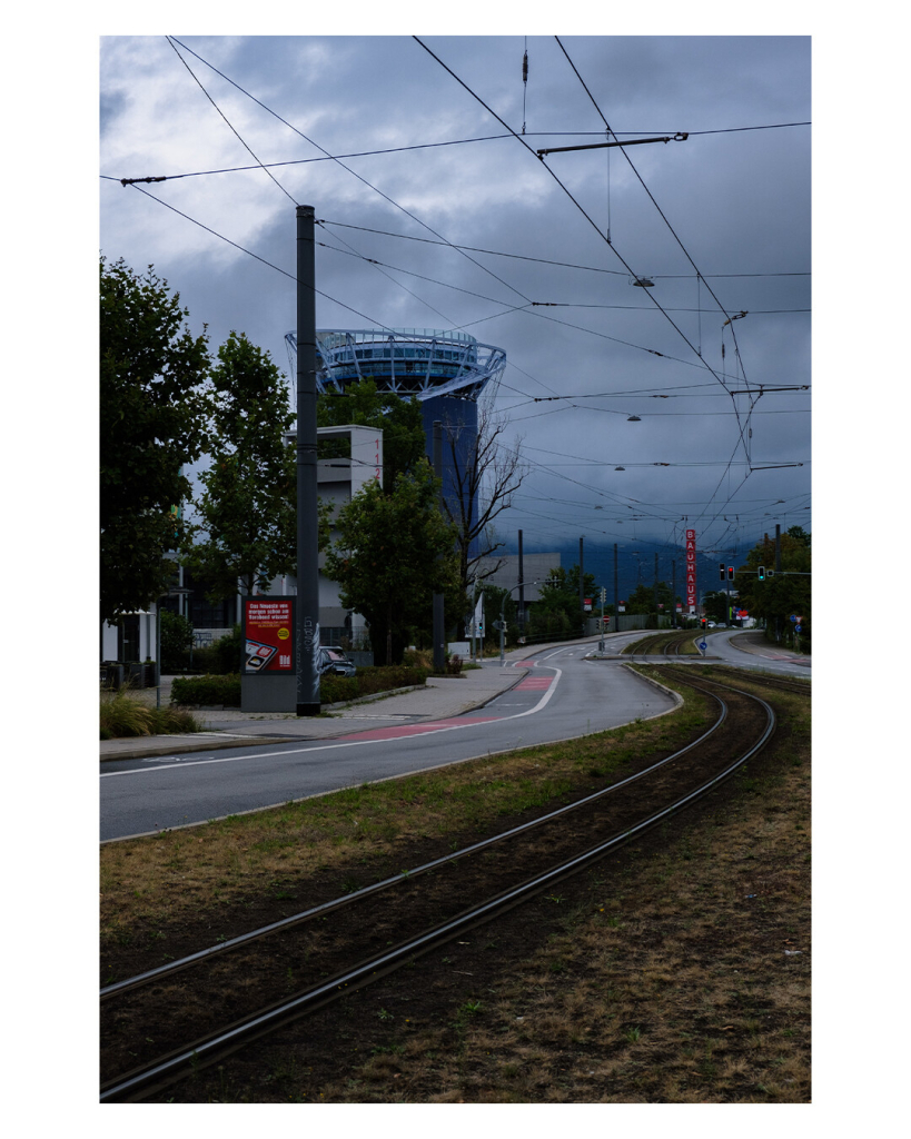 Foto im Hochformat. Schienen verlaufen vor n der linken unteren Ecke Richtung Horizont. Links und rechts daneben ist eine Straße. Am Ende der Schienen erhebt sich ein roter Turm aus Klötzen, auf dem Bauhaus steht. Der Himmel ist düster und bewölkt. 