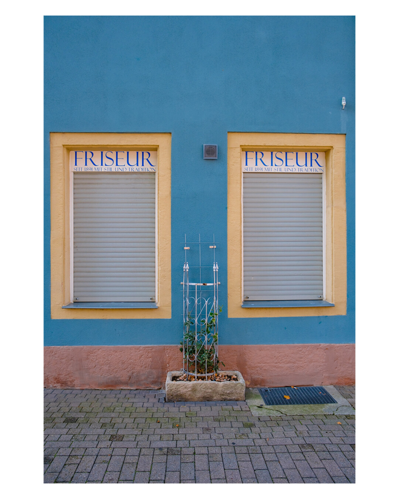 Foto im Hochformat. Eine blaue Hauswand. Nebeneinander sind zwei gleiche rechteckige Fenster, gelb umrahmt. Die Rollläden sind unten, am oberen Ende steht auf weißem Untergrund „Friseur seit 1891 mit Stil und Tradition“. Der untere Teil des Hauswand ist altrosa. Zwischen den Fenstern ist ein kleines mit Stein umrandetes Blumenbeet, darin ist eine runde Metallkonstruktion, an der eine Pflanze empor wächst. Der Boden vor der Hauswand ist Kopfsteinpflaster. Alles im Bild ist sehr symmetrisch. 