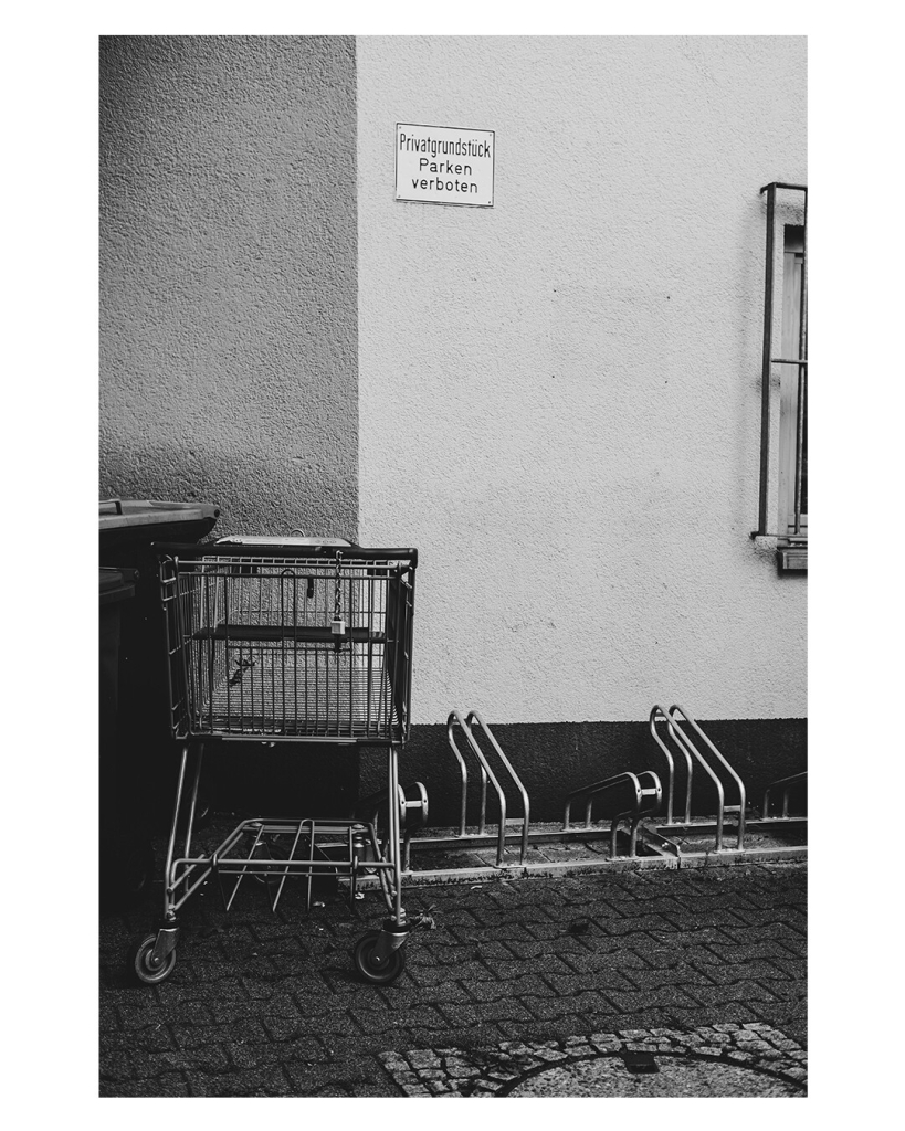 Schwarz weiß Foto im Hochformat. An einer Hausecke steht ein Einkaufswagen und ein Fahrradständer. An der Hauswand hängt ein Schild auf dem steht „Privatgrundstück parken verboten“. 