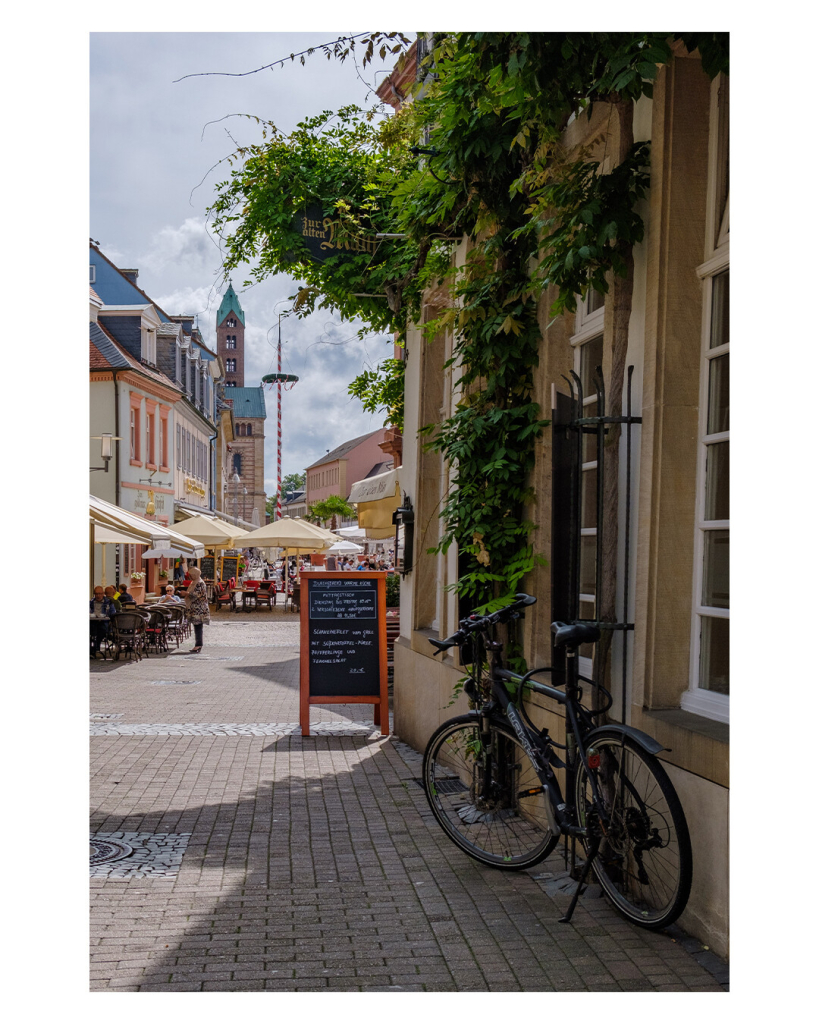 Foto im Hochformat. Eine Fußgängerzone in der Altstadt. Vorn rechts im Bild ist ein Restaurant, die Fassade ist mit viel grün begangen, davor steht ein Fahrrad und ein Schild mit den Tagesangeboten. Weiter hinten links sind weitere Cafés mit Tischen und Stühlen unter Sonnenschirmen. Am Horizont ist ein Kirche mit hohem Turm. Das Wetter ist sonnig, der Himmel leicht bewölkt. 