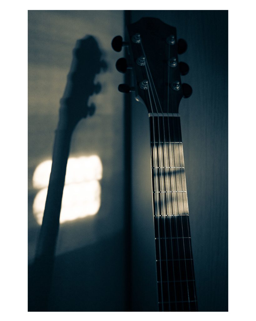 Foto im Hochformat. Der Hals einer akustischen Gitarre, die in einem dunklen Raum steht. Drei Bünde sind von der Sonne beschienen. An der wand hinter der Gitarre ist der Schatten des Halses und ein kleiner Teil wird von der Sonne beschienen. 